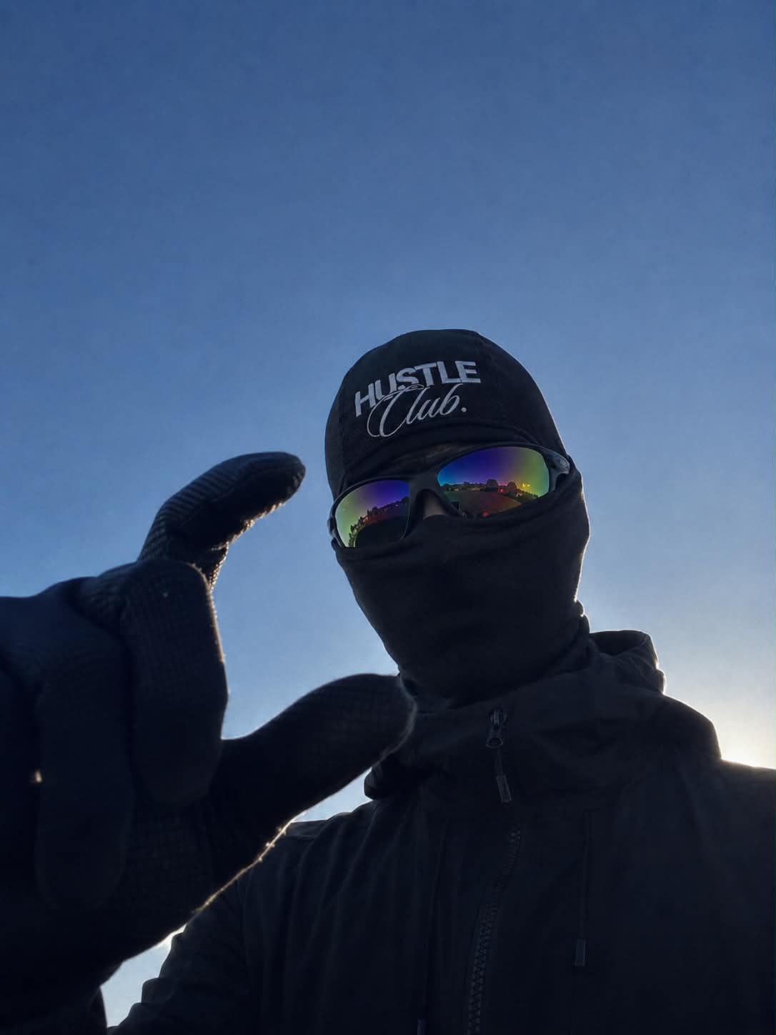 Modèle en cagoule noire et lunettes de soleil, contre-plongée sur ciel bleu.