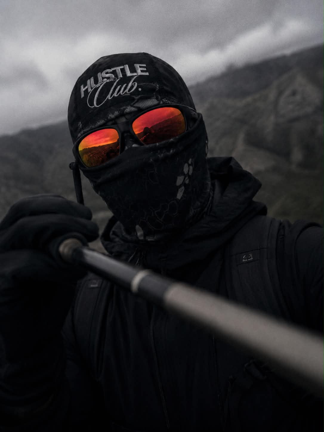 Modèle en cagoule Camouflage Python Noir avec lunettes rouges, montagne en arrière-plan.
