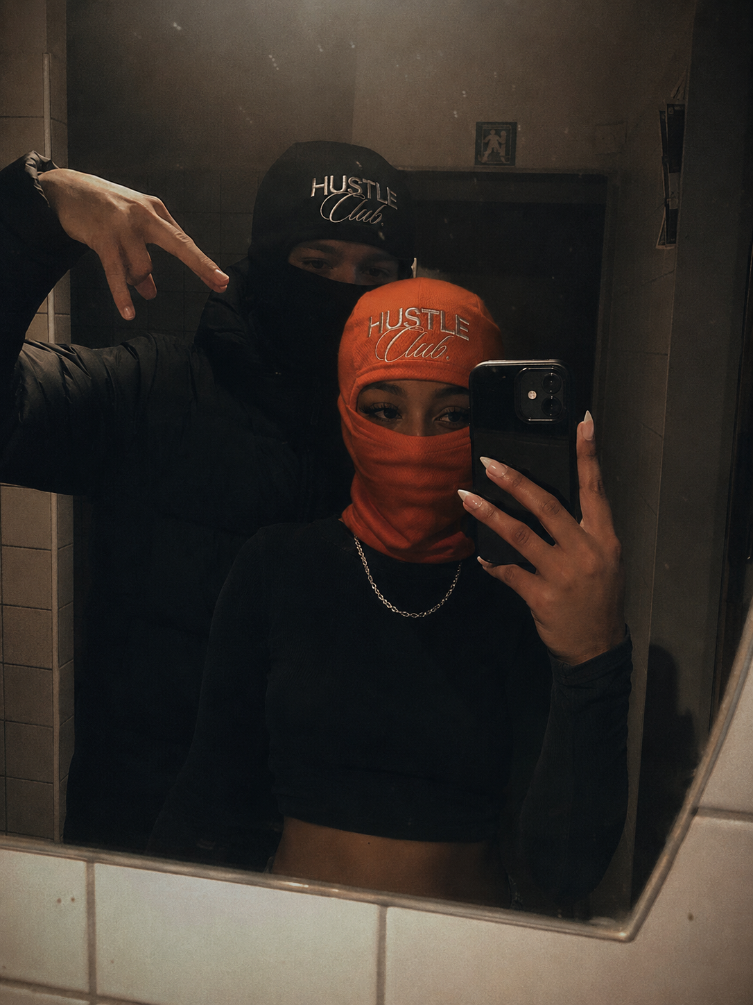 Couple en cagoules noire et orange, selfie miroir dans des toilettes carrelées.