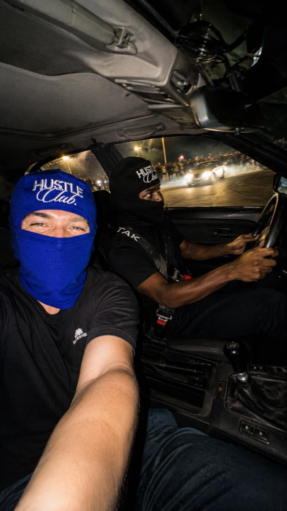 Deux modèles en cagoule bleue et noire à l'intérieur d'une voiture, car meet nocturne en arrière-plan.