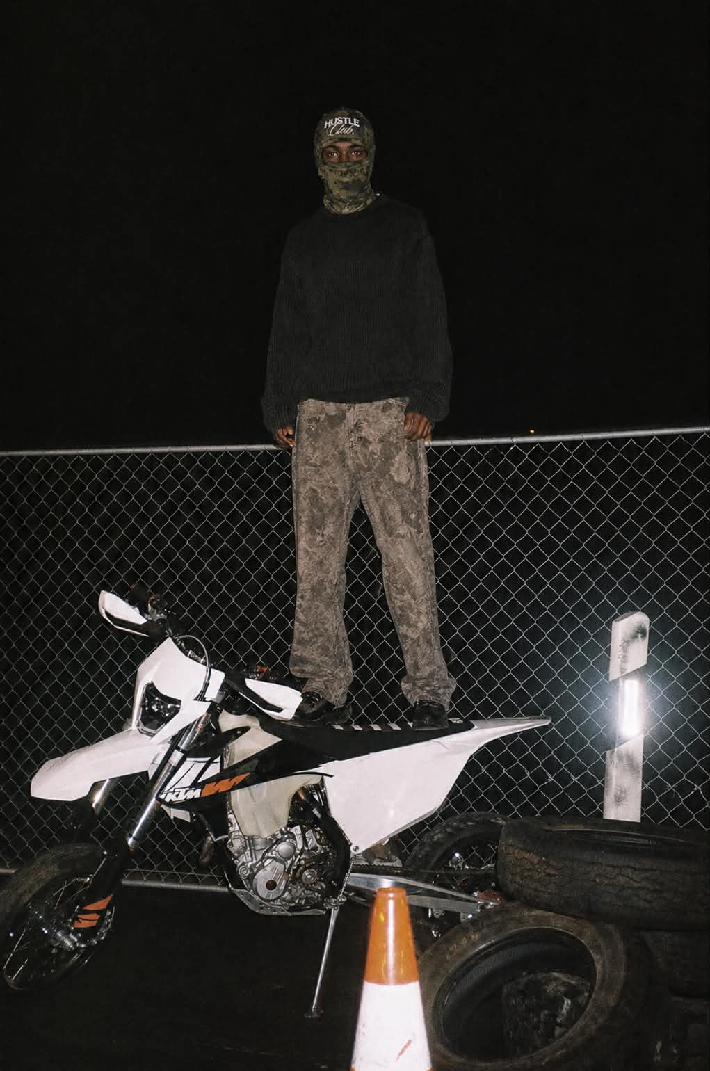 Modèle en cagoule Camouflage Vert debout sur une moto KTM blanche, la nuit.