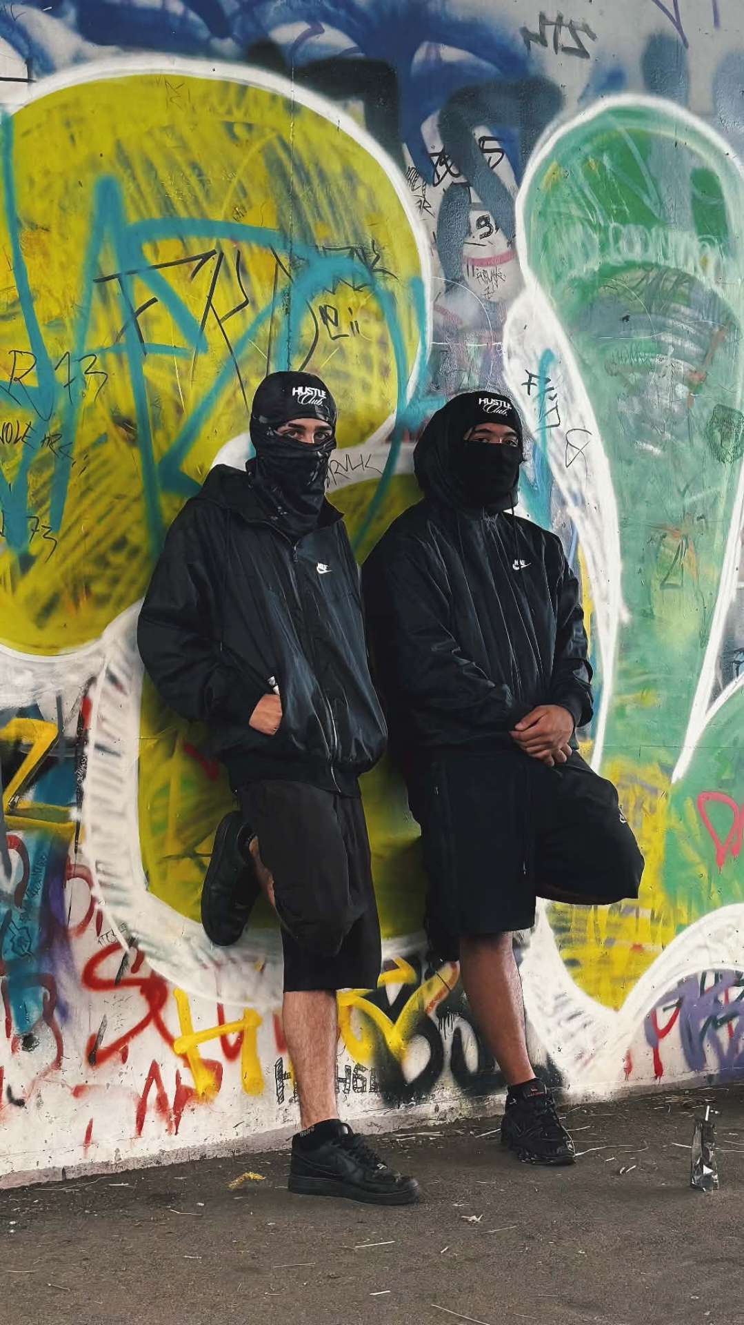 Deux modèles en cagoule noire adossés à un mur couvert de graffitis colorés.