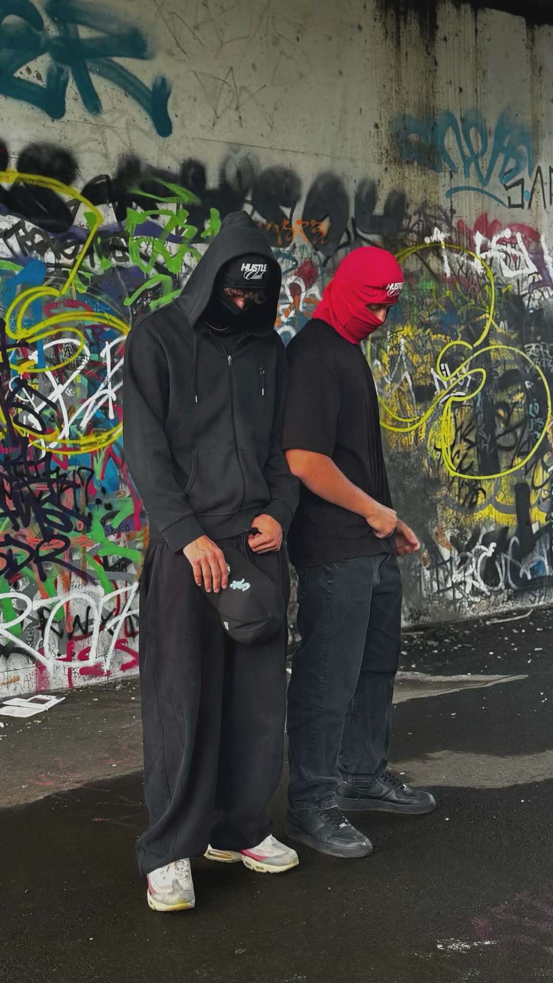 Deux modèles en cagoule noire et rouge devant un mur couvert de graffitis.