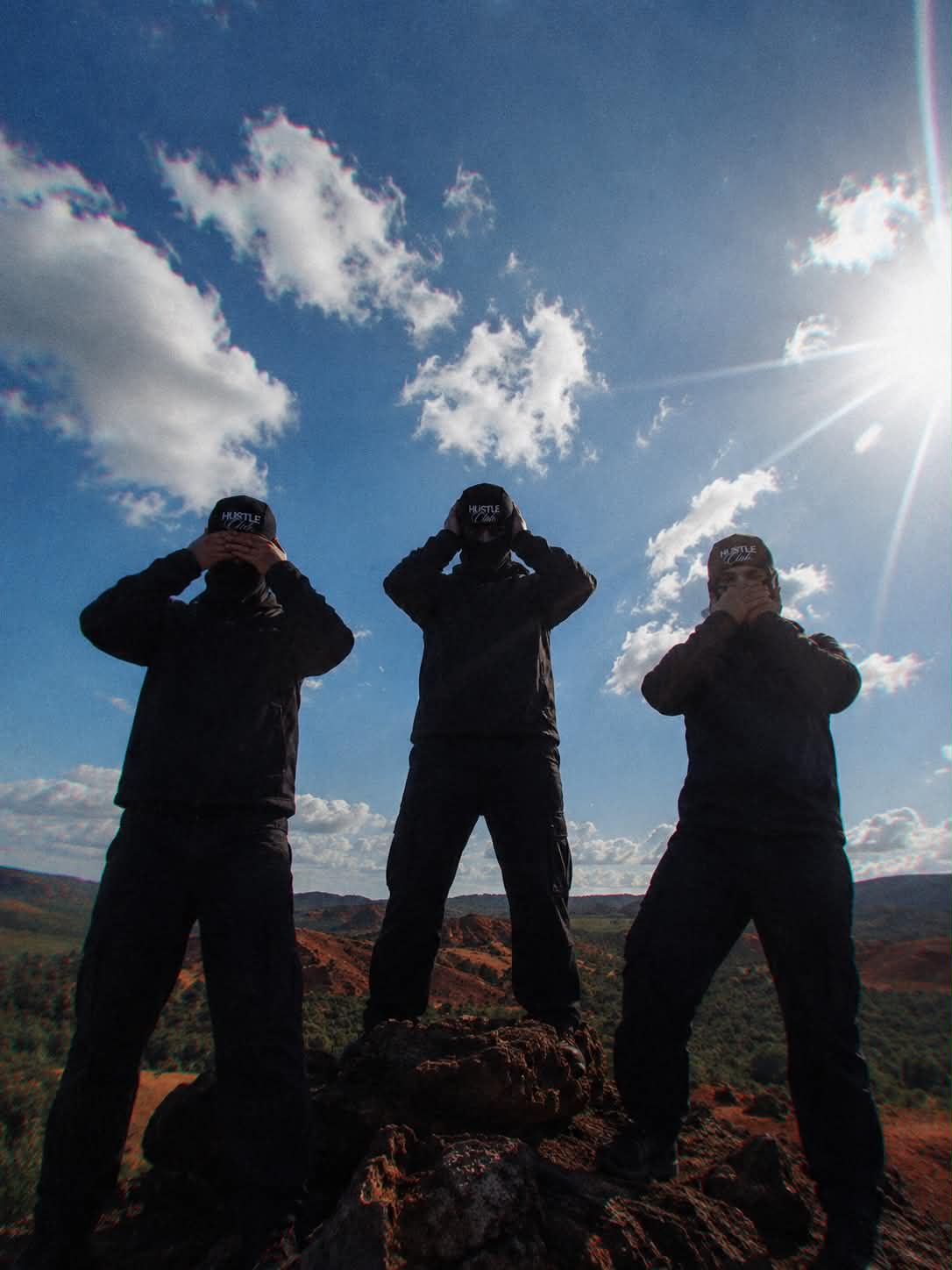 Trois silhouettes en cagoule noire face au soleil, sur un promontoire de montagne.