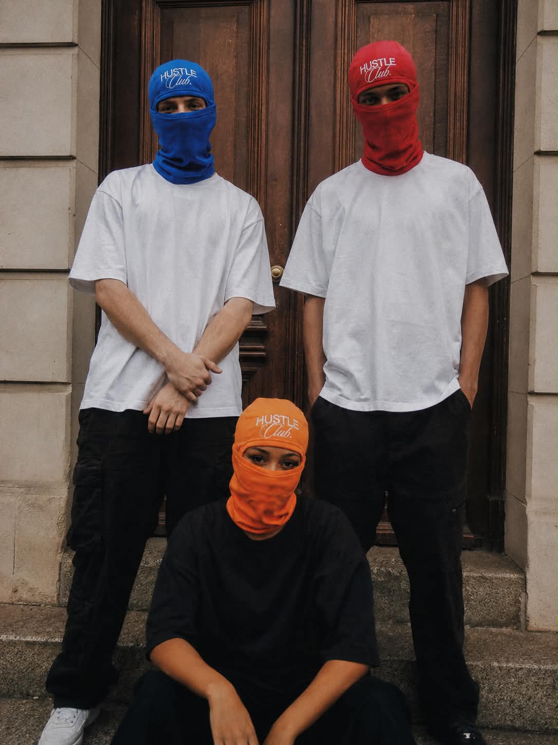 Trois modèles en cagoule bleue, rouge et orange devant une porte en bois.