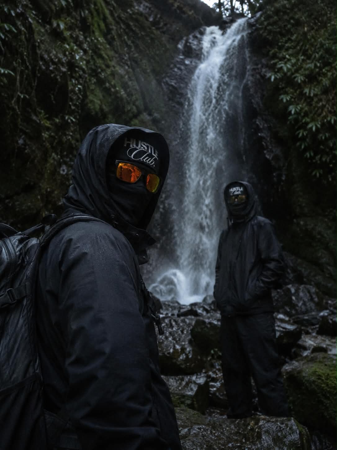 Deux modèles en cagoule noire devant une cascade en pleine forêt.