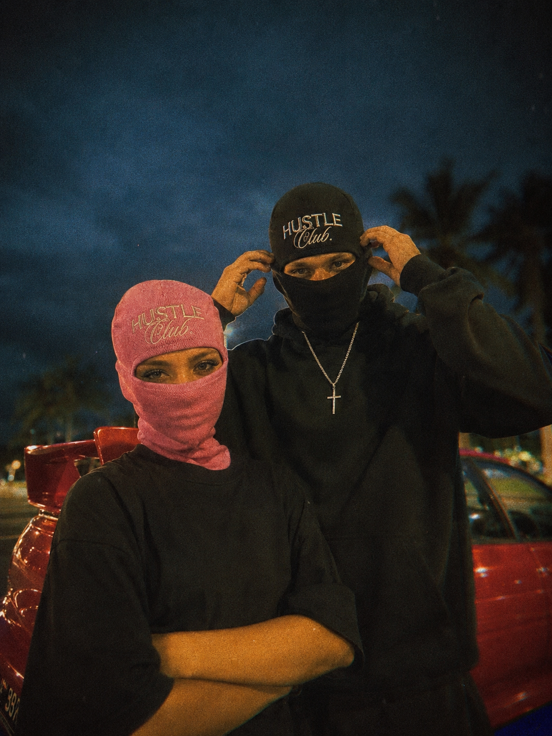 Couple en cagoules noire et rose devant une voiture rouge, ciel nocturne.